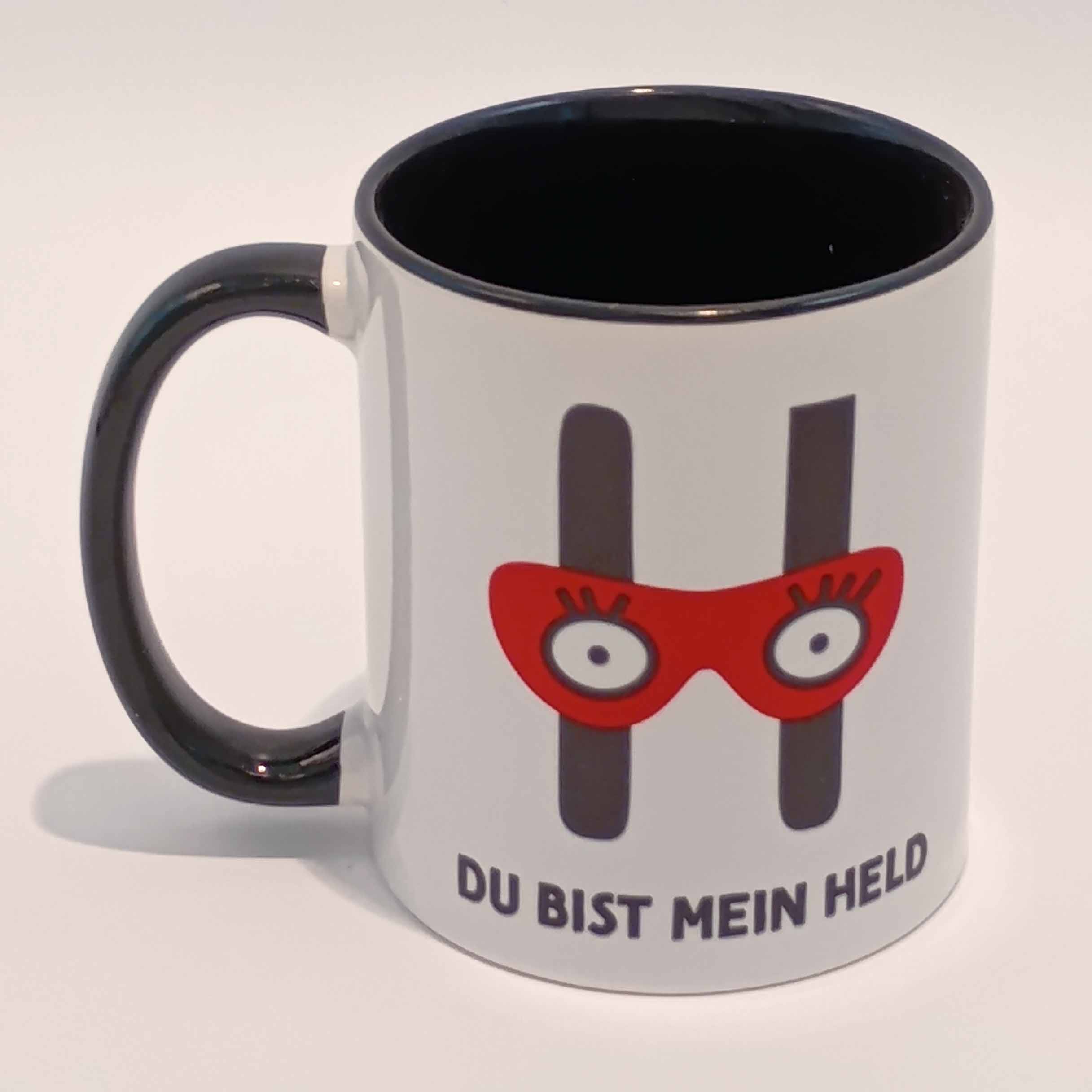 Tasse Du bist mein Held 1 Eine weiße Tasse mit schwarzem Henkel und Innenraum. Darauf das große H mit der Augenbinde vom Edition Helden Logo und der Spruch "Du bist meine Held!" in schwarzen Großbuchstaben. Das Motiv und der Spruch sind auf beiden Seiten jeweils einmal abgedruckt. Die Tasse steht auf weißem Hintergrund und der Henkel zeigt nach links.