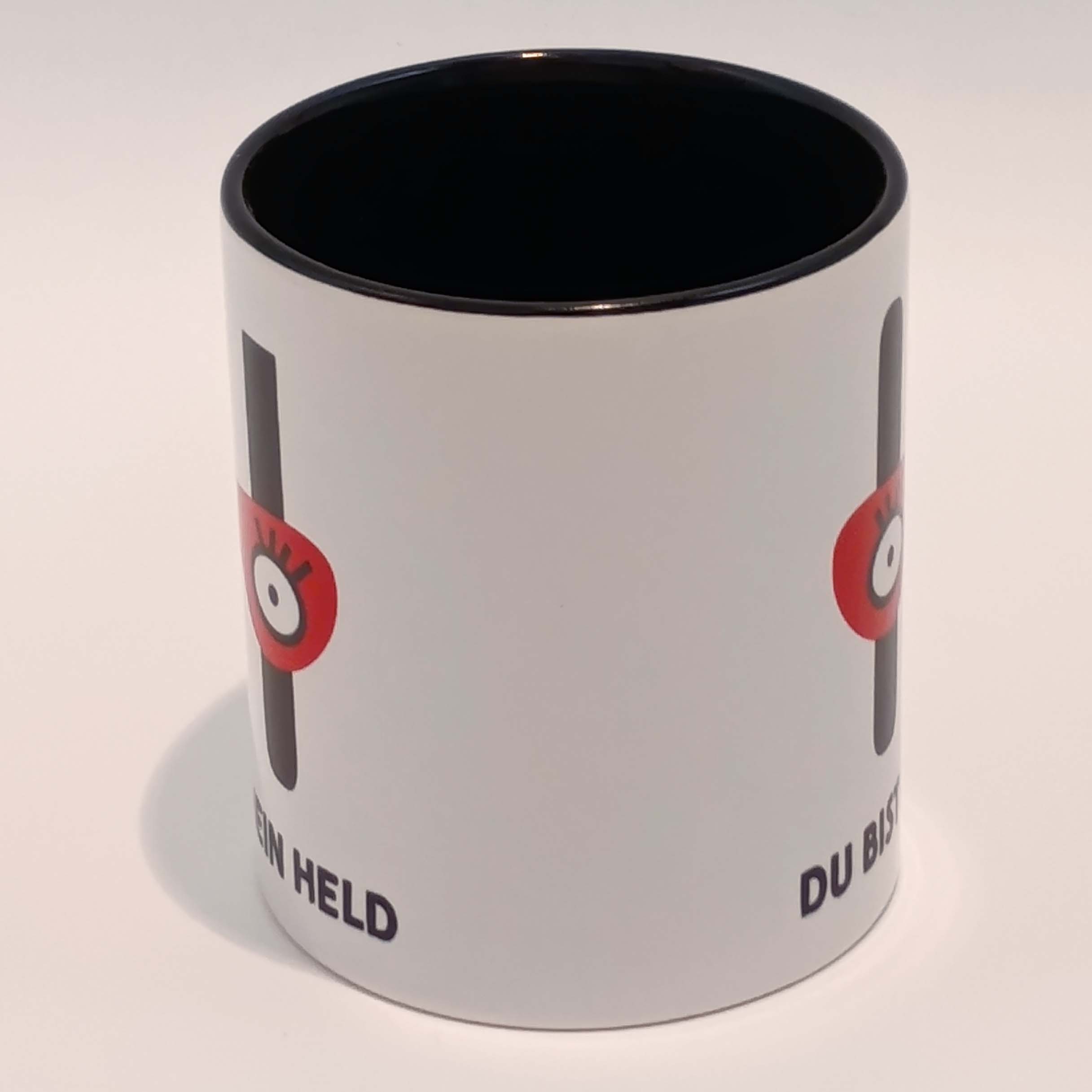 Tasse Du bist mein Held 4 Eine weiße Tasse mit schwarzem Henkel und Innenraum. Darauf das große H mit der Augenbinde vom Edition Helden Logo und der Spruch "Du bist meine Held!" in schwarzen Großbuchstaben. Das Motiv und der Spruch sind auf beiden Seiten jeweils einmal abgedruckt. Die Tasse steht auf weißem Hintergrund und der Henkel zeigt von uns weg.