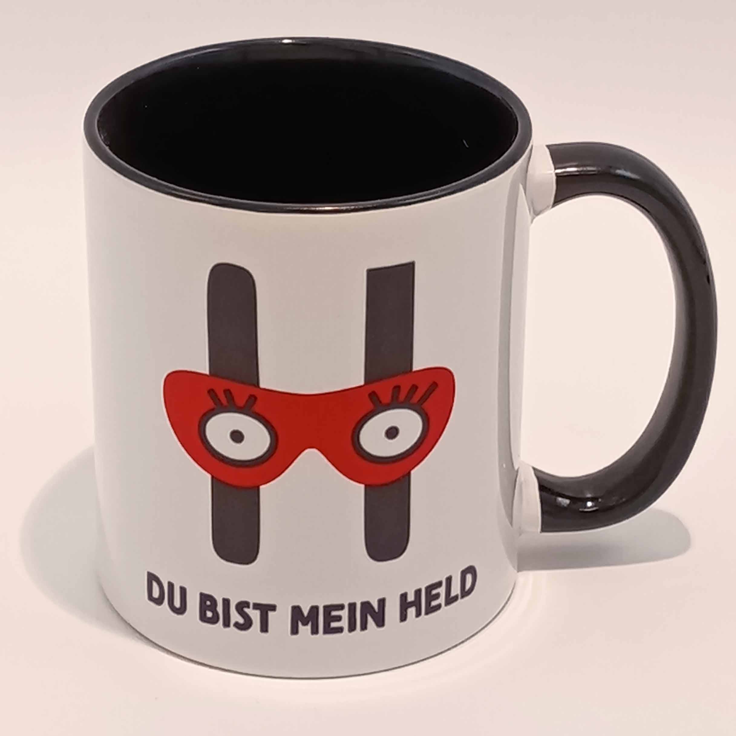 Tasse Du bist mein Held 2 Eine weiße Tasse mit schwarzem Henkel und Innenraum. Darauf das große H mit der Augenbinde vom Edition Helden Logo und der Spruch "Du bist meine Held!" in schwarzen Großbuchstaben. Das Motiv und der Spruch sind auf beiden Seiten jeweils einmal abgedruckt. Die Tasse steht auf weißem Hintergrund und der Henkel zeigt nach rechts.