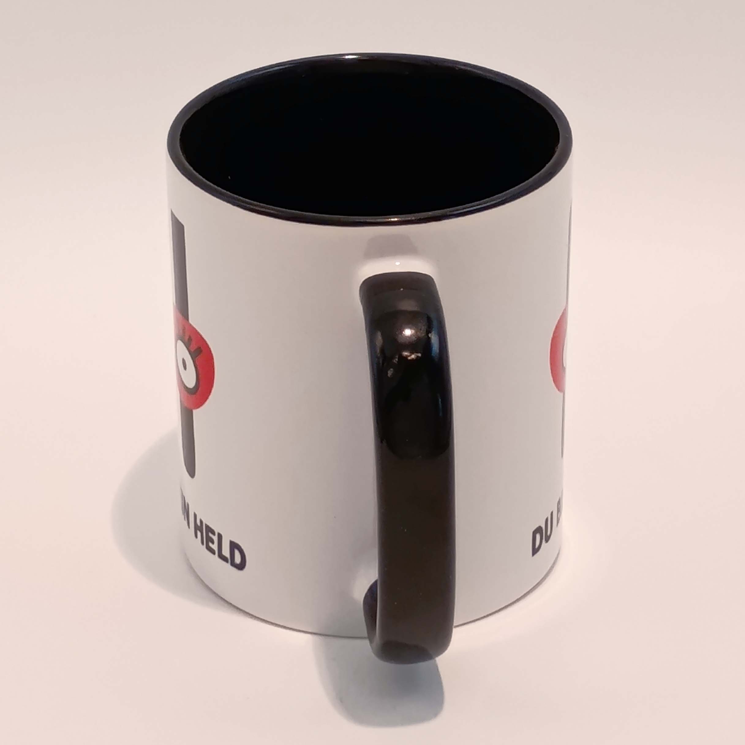 Tasse Du bist mein Held 3 Eine weiße Tasse mit schwarzem Henkel und Innenraum. Darauf das große H mit der Augenbinde vom Edition Helden Logo und der Spruch "Du bist meine Held!" in schwarzen Großbuchstaben. Das Motiv und der Spruch sind auf beiden Seiten jeweils einmal abgedruckt. Die Tasse steht auf weißem Hintergrund und der Henkel zeigt zu uns.