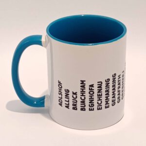 Weiße Tasse mit mittel blauem Henkel und Innenwand. Darauf sind die zum Landkreis Fürstenfeldbruck gehörenden Ortsnamen in lokalem Dialekt abgedruckt. Die Wörter sind schwarz und in Großbuchstaben. Sie sind vertikal einzeln nebeneinander gedruckt und bilden eine lange Zeile, die die Tasse umläuft. Die Zeile hält wenige Zentimeter Abstand zum Henkel. Die geschriebenen Wörter sind schwarz. Die Tasse steht auf einem weißen Hintergrund. Der Henkel zeigt nach links.