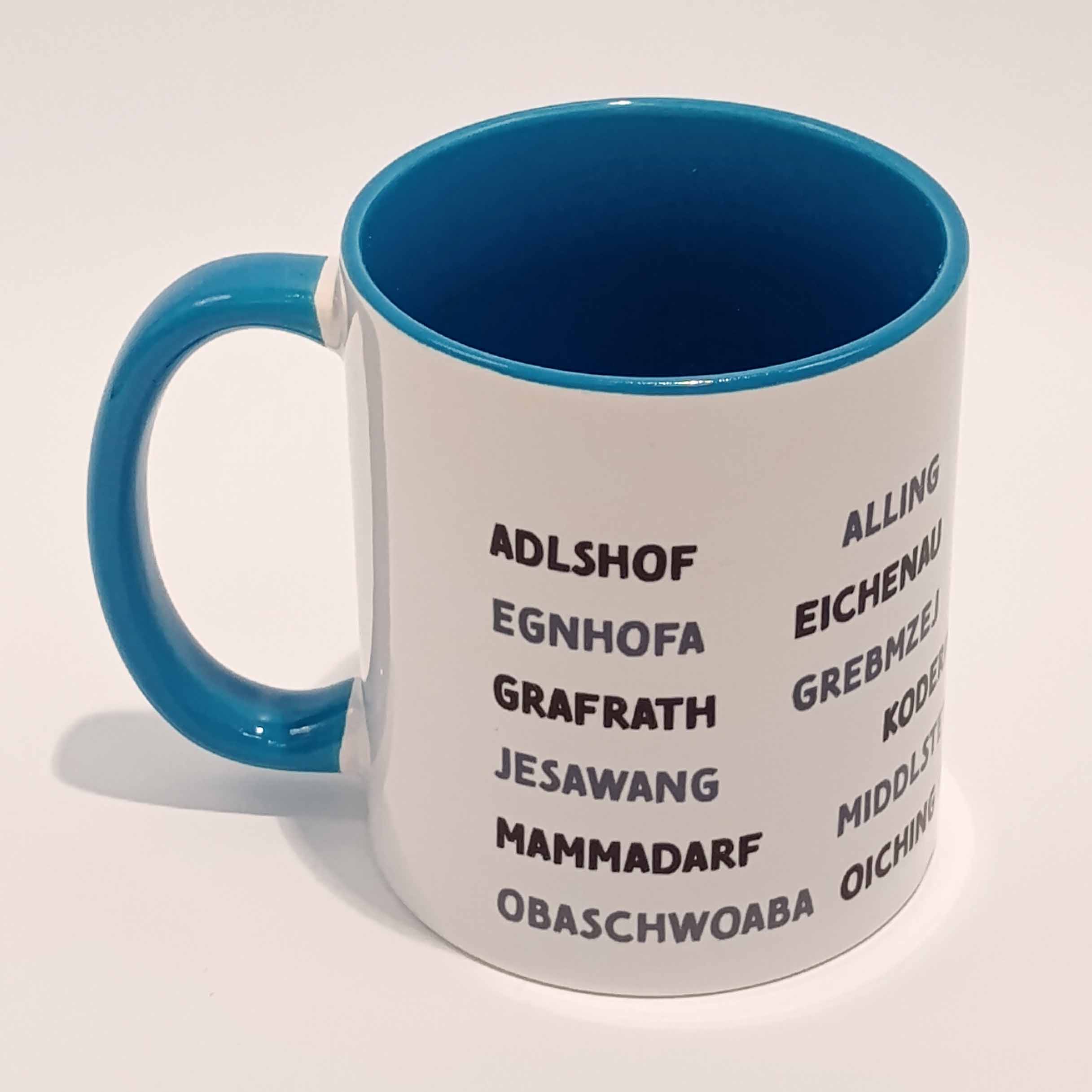 Tasse Landkreis Bruck blau 1 Weiße Tasse mit mittel blauem Henkel und Innenwand. Darauf sind die zum Landkreis Fürstenfeldbruck gehörenden Ortsnamen in lokalem Dialekt abgedruckt. Sie sind in Großbuchstaben gedruckt und bilden sechs horizontal laufende Zeilen, die mit einigen Zentimetern Abstand vom Henkel anfangen als auch aufhören. Die geschriebenen Wörter variieren zwischen schwarz und einem mitteldunklen grau. Die Tasse steht auf einem weißen Hintergrund. Der Henkel zeigt nach links.