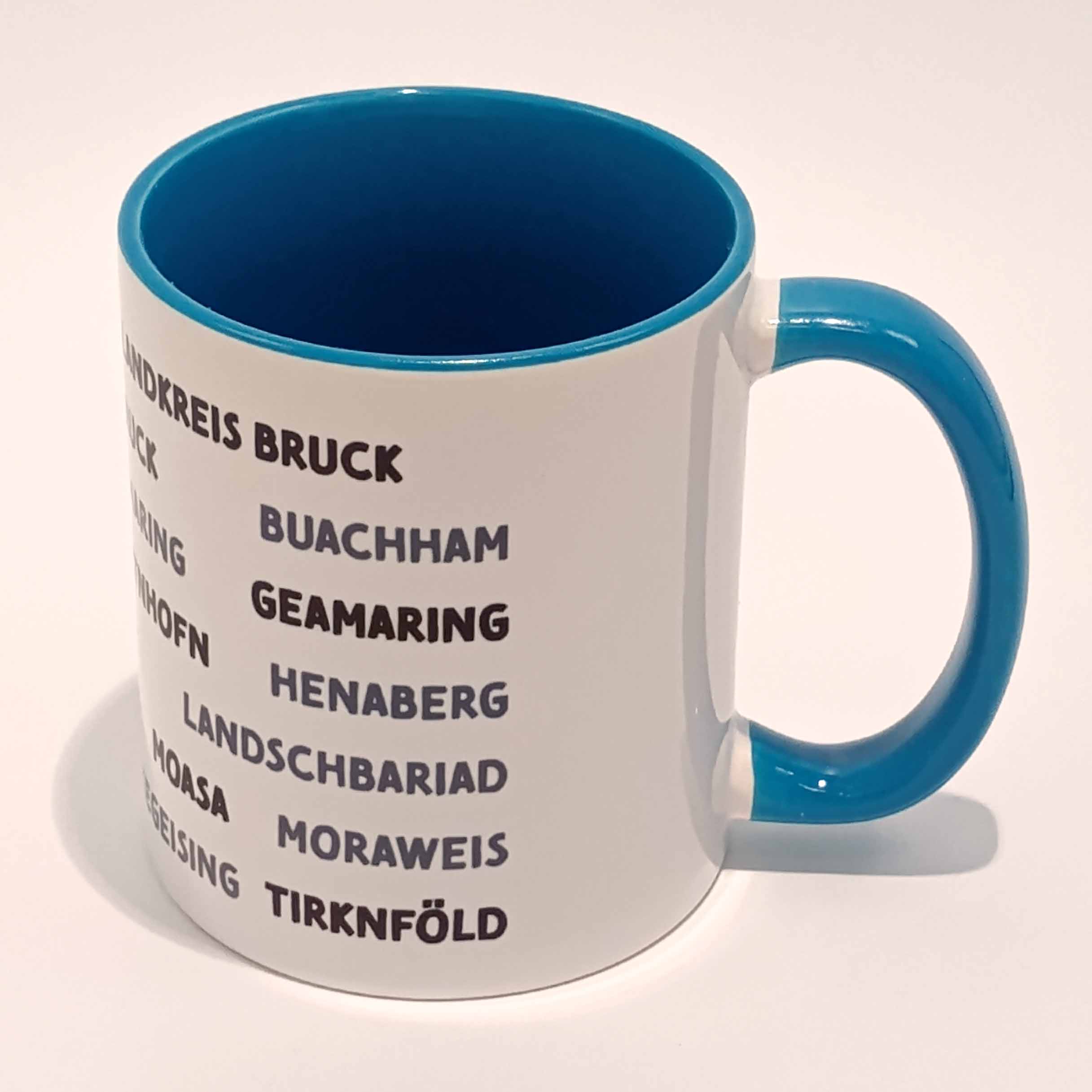 Tasse Landkreis Bruck blau 2 Weiße Tasse mit mittel blauem Henkel und Innenwand. Darauf sind die zum Landkreis Fürstenfeldbruck gehörenden Ortsnamen in lokalem Dialekt abgedruckt. Sie sind in Großbuchstaben gedruckt und bilden sechs horizontal laufende Zeilen, die mit einigen Zentimetern Abstand vom Henkel anfangen als auch aufhören. Die geschriebenen Wörter variieren zwischen schwarz und einem mitteldunklen grau. Die Tasse steht auf einem weißen Hintergrund. Der Henkel zeigt nach rechts.