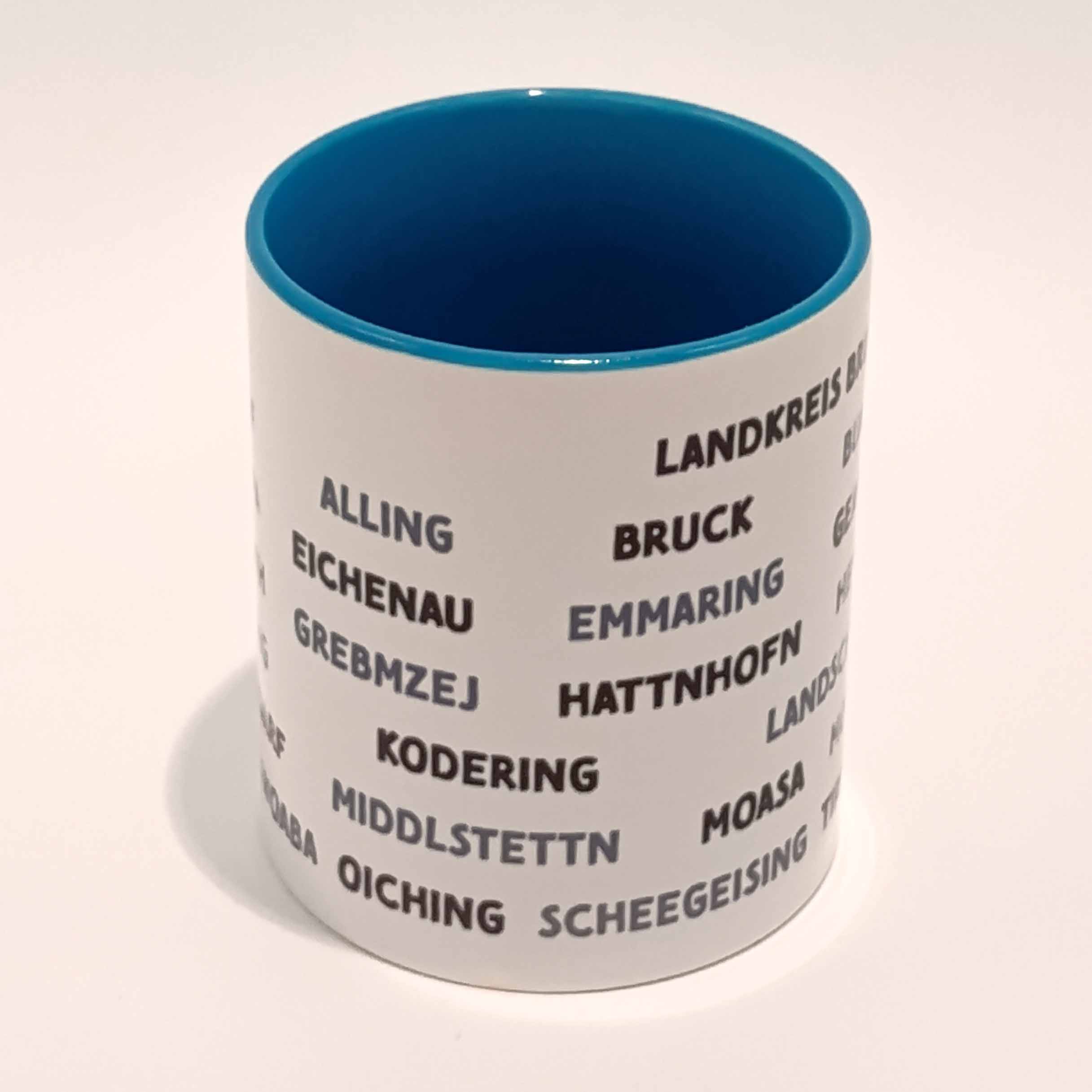 Tasse Landkreis Bruck blau 3 Weiße Tasse mit mittel blauem Henkel und Innenwand. Darauf sind die zum Landkreis Fürstenfeldbruck gehörenden Ortsnamen in lokalem Dialekt abgedruckt. Sie sind in Großbuchstaben gedruckt und bilden sechs horizontal laufende Zeilen, die mit einigen Zentimetern Abstand vom Henkel anfangen als auch aufhören. Die geschriebenen Wörter variieren zwischen schwarz und einem mitteldunklen grau. Die Tasse steht auf einem weißen Hintergrund. Der Henkel ist von uns weg gedreht.
