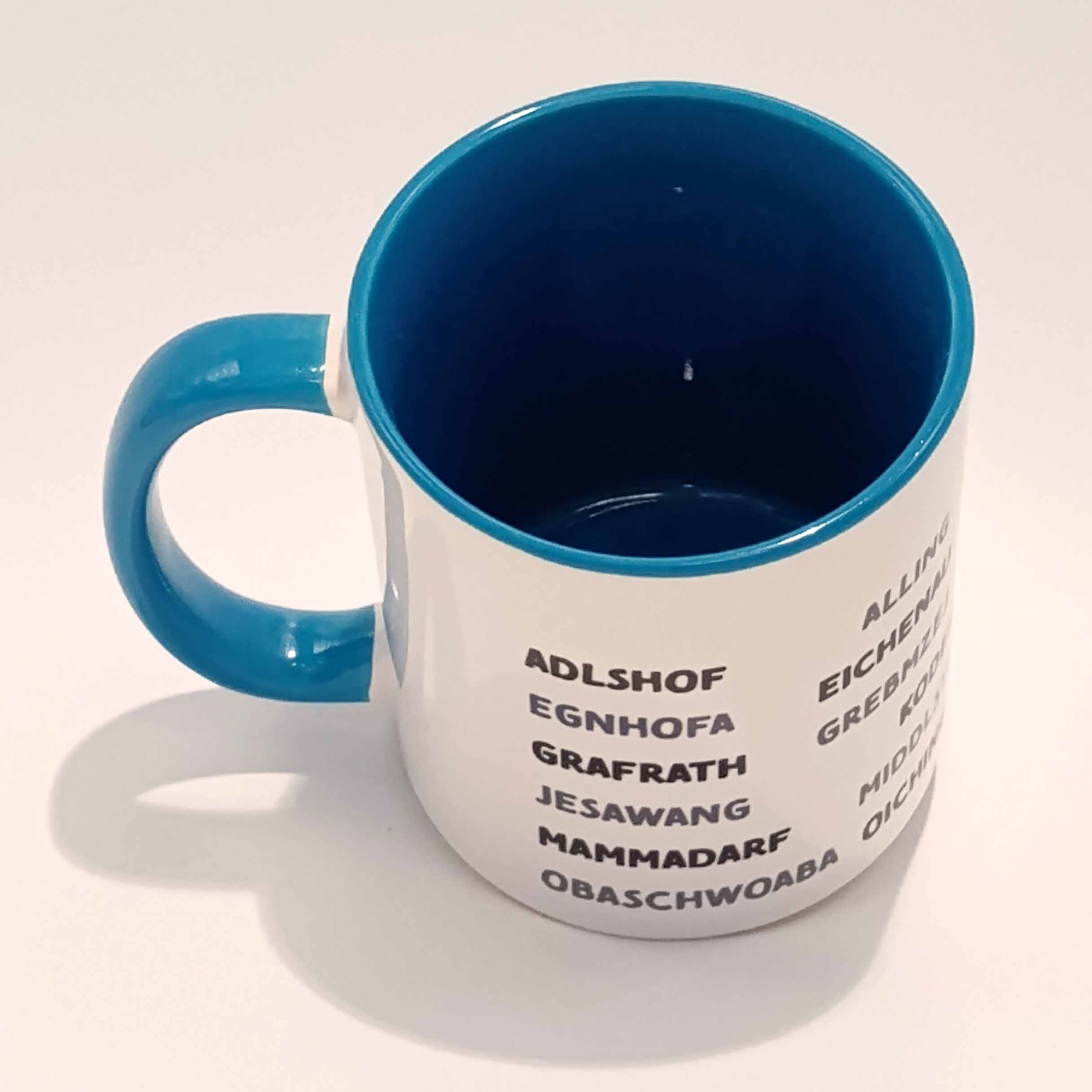 Tasse Landkreis Bruck blau 4 Weiße Tasse mit mittel blauem Henkel und Innenwand. Darauf sind die zum Landkreis Fürstenfeldbruck gehörenden Ortsnamen in lokalem Dialekt abgedruckt. Sie sind in Großbuchstaben gedruckt und bilden sechs horizontal laufende Zeilen, die mit einigen Zentimetern Abstand vom Henkel anfangen als auch aufhören. Die geschriebenen Wörter variieren zwischen schwarz und einem mitteldunklen grau. Die Tasse steht auf einem weißen Hintergrund. Der Henkel zeigt nach links und wir blicken von leicht schräg oben in die Tasse hinein.