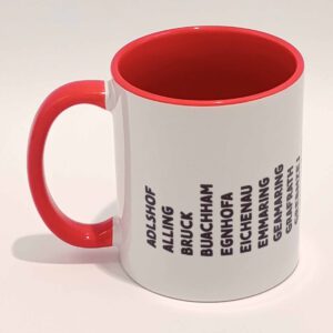 Weiße Tasse mit grell rotem Henkel und Innenwand. Darauf sind die zum Landkreis Fürstenfeldbruck gehörenden Ortsnamen in lokalem Dialekt abgedruckt. Die Wörter sind schwarz und in Großbuchstaben. Sie sind vertikal einzeln nebeneinander gedruckt und bilden eine lange Zeile, die die Tasse umläuft. Die Zeile hält wenige Zentimeter Abstand zum Henkel. Die geschriebenen Wörter sind schwarz. Die Tasse steht auf einem weißen Hintergrund. Der Henkel zeigt nach links.