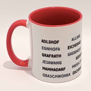 Weiße Tasse mit grell rotem Henkel und Innenwand. Darauf sind die zum Landkreis Fürstenfeldbruck gehörenden Ortsnamen in lokalem Dialekt und Großbuchstaben abgedruckt. Diese bilden sechs horizontal laufende Zeilen, die mit einigen Zentimetern Abstand vom Henkel anfangen als auch aufhören. Die geschriebenen Wörter variieren zwischen schwarz und einem mitteldunklen grau. Die Tasse steht auf einem weißen Hintergrund. Der Henkel zeigt nach links.